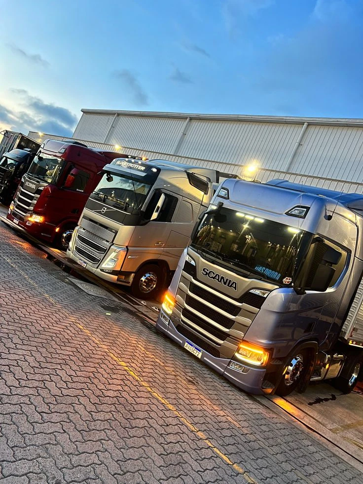 Três caminhões cavalo-mecânico de cores diferentes parados lado a lado no pátio de uma transportadora, ao entardecer